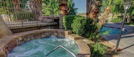 A hot tub surrounded by stone walls and palm trees, with a pool and lounge chairs in the background.