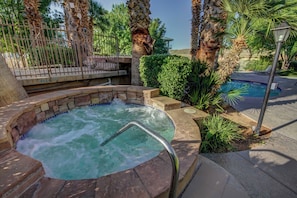 A hot tub surrounded by stone walls and palm trees, with a pool and lounge chairs in the background.