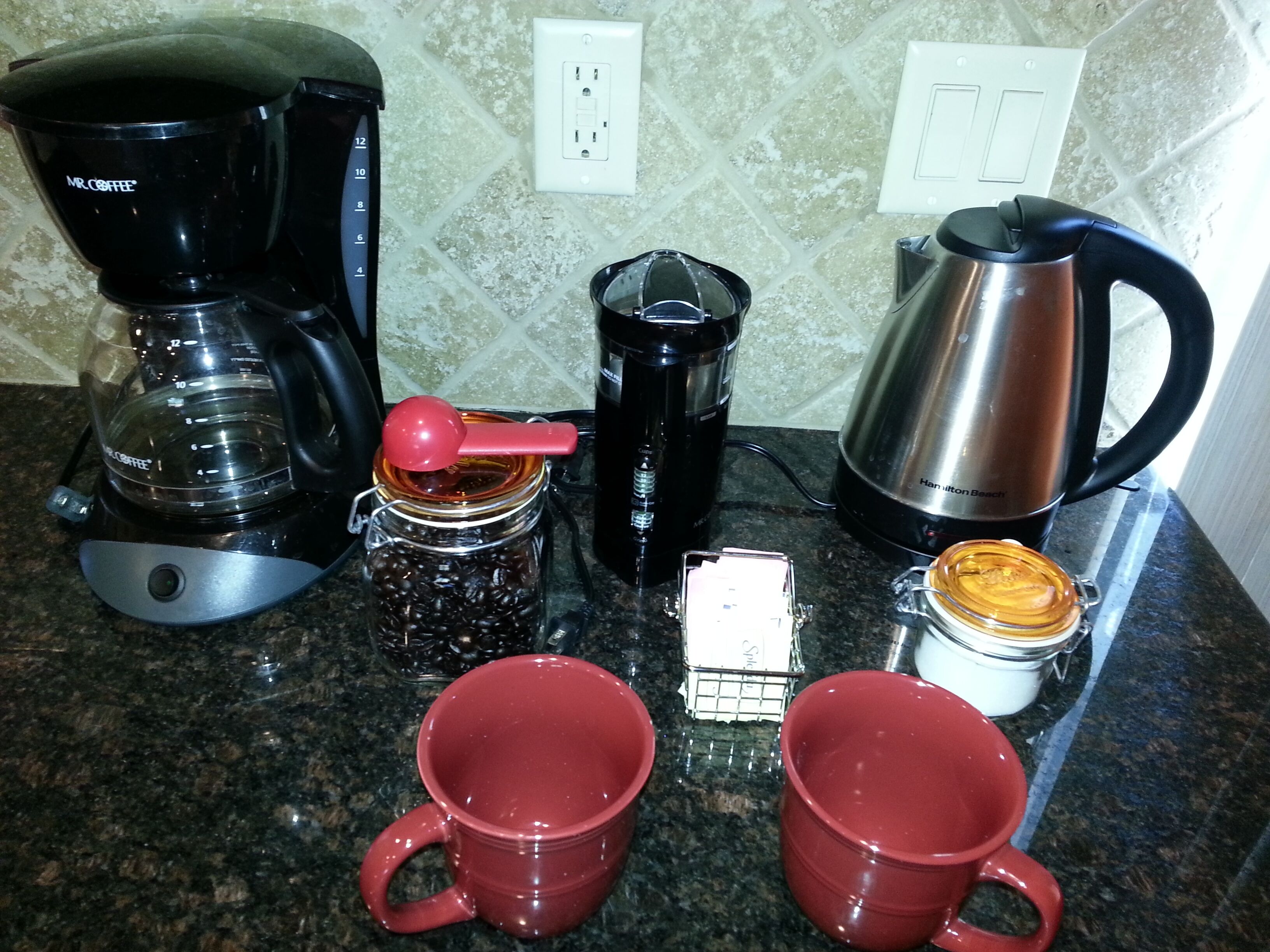Una encimera de cocina con una cafetera, un hervidor, un tarro con una cuchara roja y dos tazas rojas.