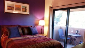 Une chambre d’hôtel comprenant un lit, une table de chevet, une lampe, un balcon avec une table et des chaises, et un tableau encadré au mur.