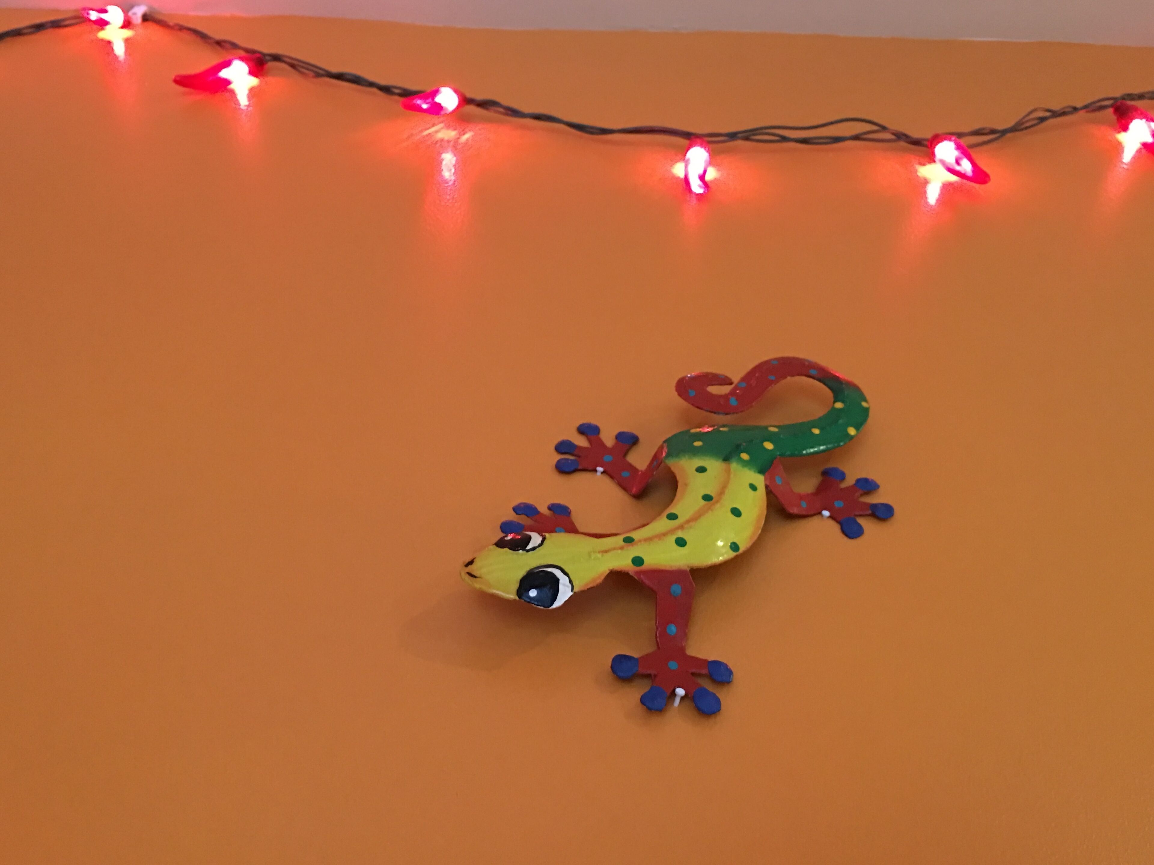Une décoration représentant un lézard aux couleurs vives, placée sur un fond orange uni, avec des guirlandes lumineuses en arrière-plan.
