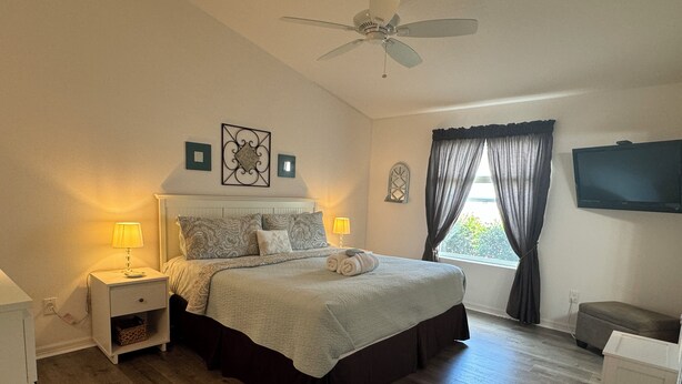 Quarto com cama, abajures, ventilador de teto, televisão e janela com cortinas.