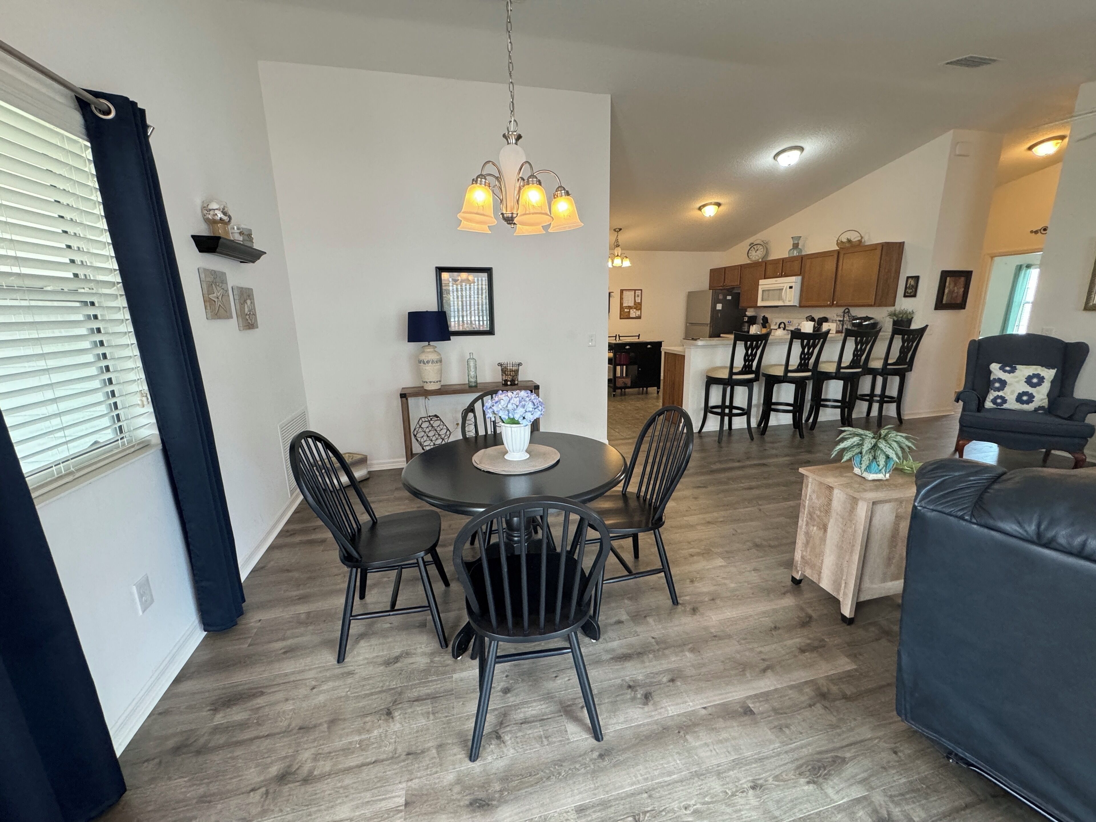 A dining area with a round table, black chairs, and a chandelier. There is a kitchen in the background with wooden cabinets and a microwave.