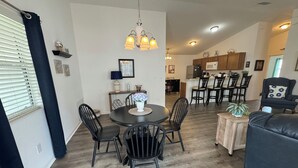 Uma sala de jantar com uma mesa redonda, cadeiras pretas e um candelabro. Ao fundo, cozinha com armários de madeira e micro-ondas.