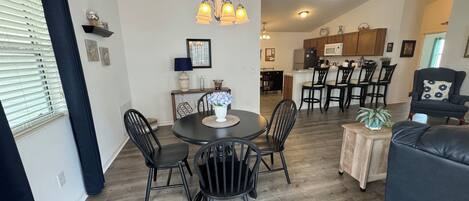 A dining area with a round table, black chairs, and a chandelier. There is a kitchen in the background with wooden cabinets and a microwave.