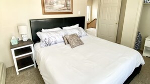 Uma cama bem arrumada com roupa de cama branca, cabeceira preta e almofadas decorativas. Há uma mesa de cabeceira com uma luminária e um quadro na parede.