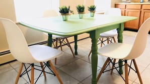 Uma sala de jantar com mesa verde e cadeiras brancas, plantas em vasos sobre a mesa e uma cozinha ao fundo.