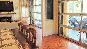 A living room with a wooden dining table, chairs, a sofa, a fireplace, and large windows. - Beautifully decorated 3bdrm/3bath near beach; unique porch with garage doors! (Folly Beach)
