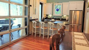 A modern kitchen with white cabinets, a stainless steel refrigerator, and a dining area with wooden chairs and a table. - Beautifully decorated 3bdrm/3bath near beach; unique porch with garage doors! (Folly Beach)