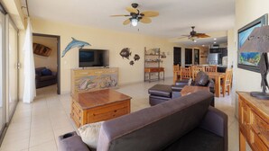 A spacious living room with a sofa, wooden coffee table, and a television. There are ceiling fans, a dining area with chairs, and a kitchen in the background.