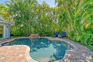A swimming pool with a curved edge, surrounded by a brick patio and lush tropical foliage.