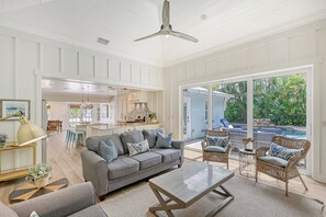 A spacious living room with a grey sofa, a coffee table, and wicker chairs. A kitchen is visible in the background, and there is a large sliding glass door leading to a pool area. - Unwind Sunshine | Heated Pool | 1.5 Blocks to Beach | Complimentary Bikes (Holmes Beach)