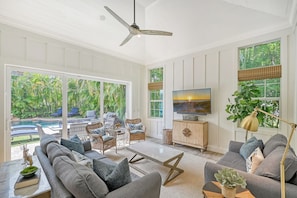 A modern living room with a large TV, comfortable seating, and a view of a pool.