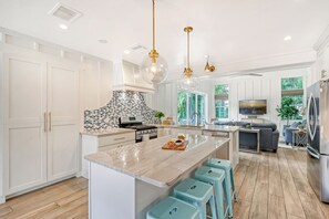 A modern kitchen with white cabinetry, a central island with stools, a stainless steel refrigerator, and a large window with a view of greenery. - Unwind Sunshine | Heated Pool | 1.5 Blocks to Beach | Complimentary Bikes (Holmes Beach)