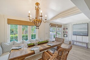 A dining room with a long table set for six, a chandelier, and large windows with blinds.