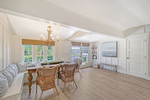 A dining area with a table set for four, surrounded by wicker chairs, a chandelier, and large windows with blinds.