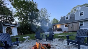 Een achtertuin met een vuurkuil, omringd door Adirondack-stoelen, een barbecue en een tuinhuis.