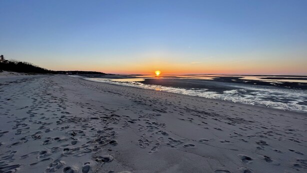 Een strand met voetafdrukken die naar het water leiden, een ondergaande zon op de achtergrond en een heldere lucht.
