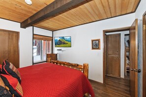 Une chambre avec un plafond en bois, un lit recouvert d’une couette rouge, un téléviseur fixé au mur et une porte donnant sur une autre pièce.