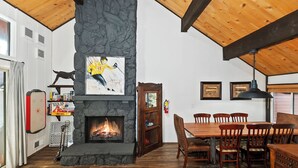 A cozy room with a stone fireplace, wooden ceiling beams, and a dining area with wooden chairs and a table. - Located at the Base of Snow Summit! Walk to Hiking Trails! Mini Air Hockey Table! (Big Bear Lake)