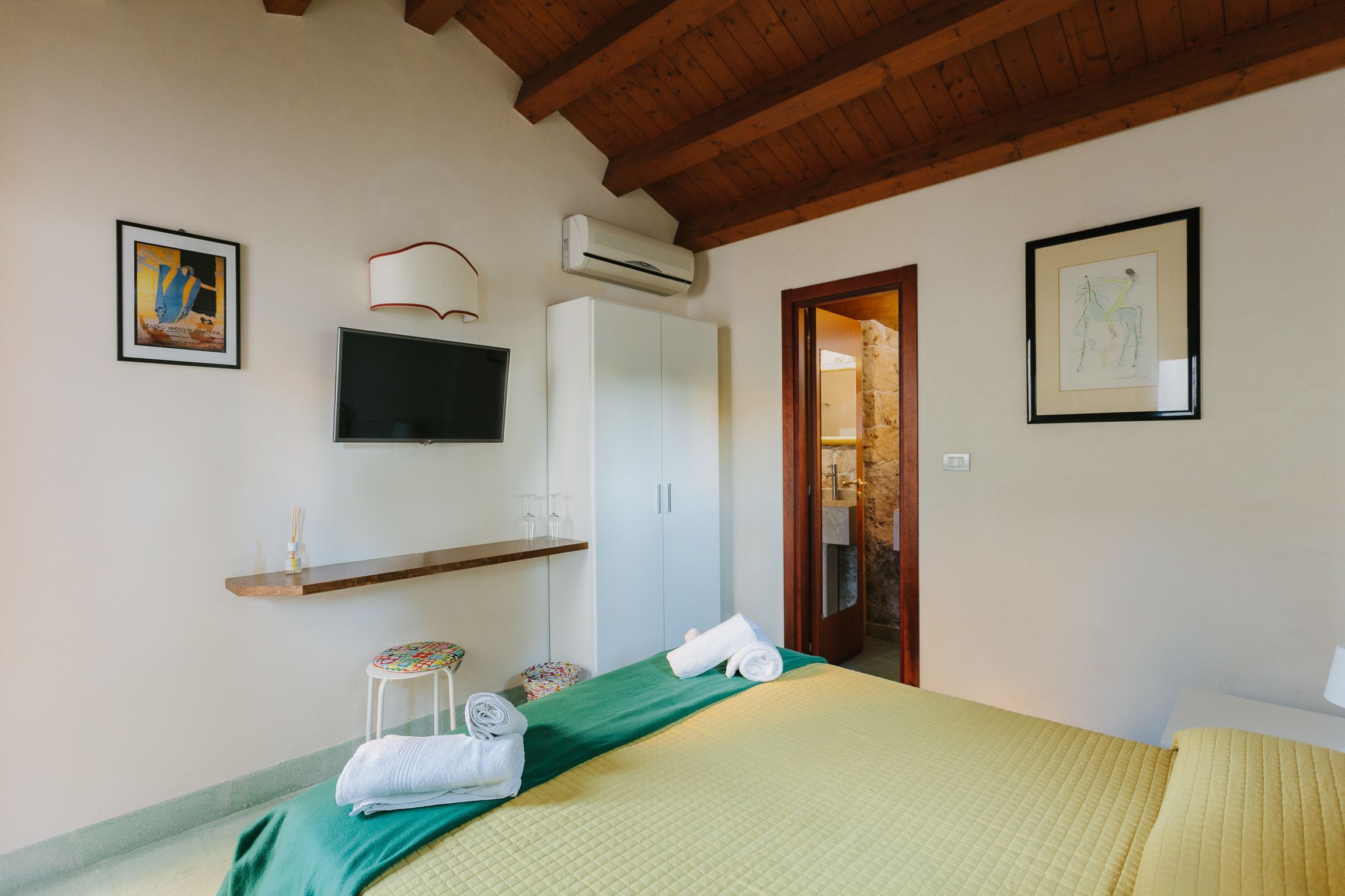 A bedroom with a bed, a TV, an air conditioner, a wooden ceiling, and artwork on the walls.