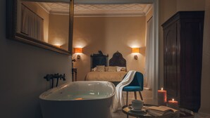 Een badkamer met een vrijstaand bad, een spiegel, een bed met twee bedlampjes, een blauwe stoel en een tafeltje met een kopje en een boek.