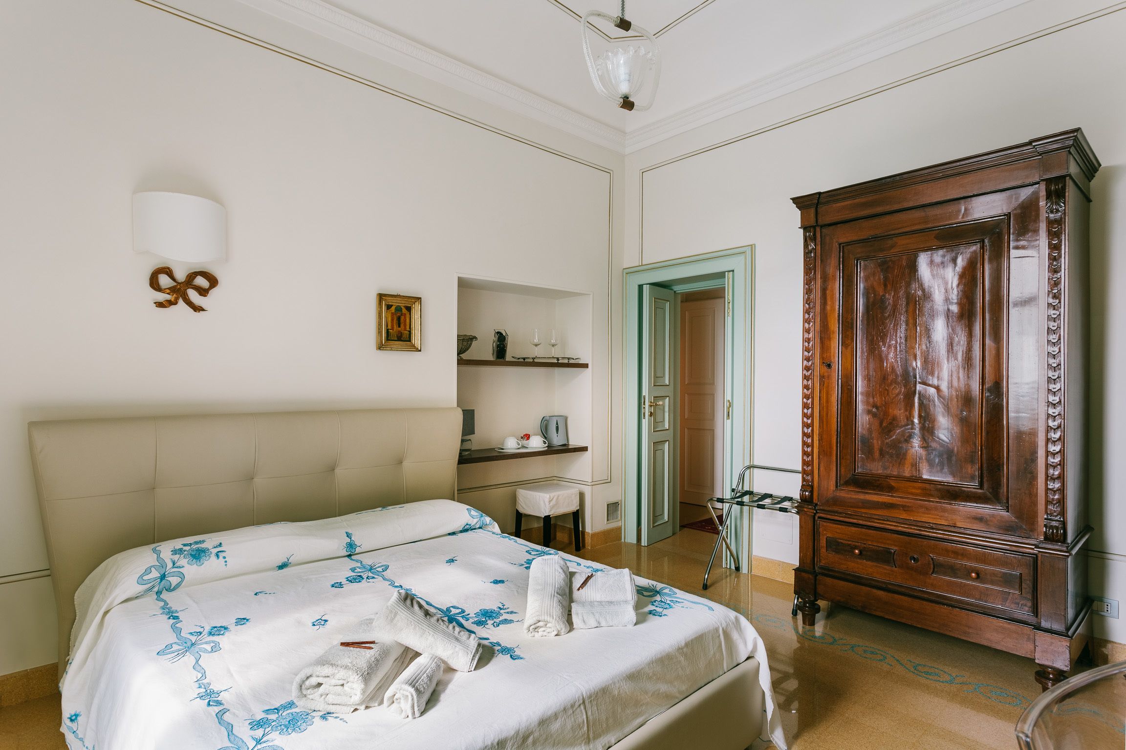 A bedroom with a bed, a wooden wardrobe, a small table, and a wall-mounted light fixture.