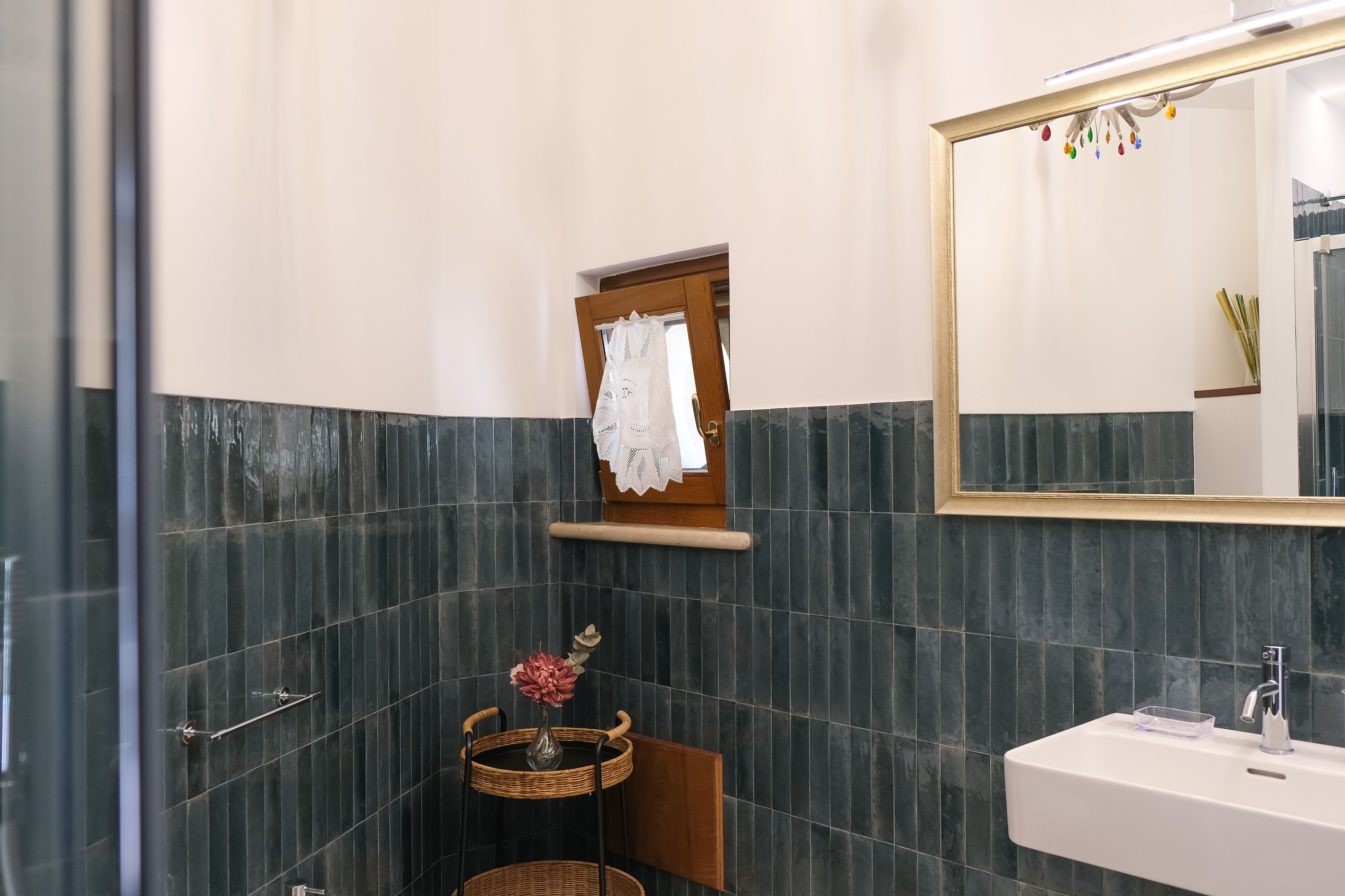 Une salle de bain avec des murs recouverts de carreaux foncés, un lavabo blanc, un miroir et une petite fenêtre ornée d’un rideau en dentelle.