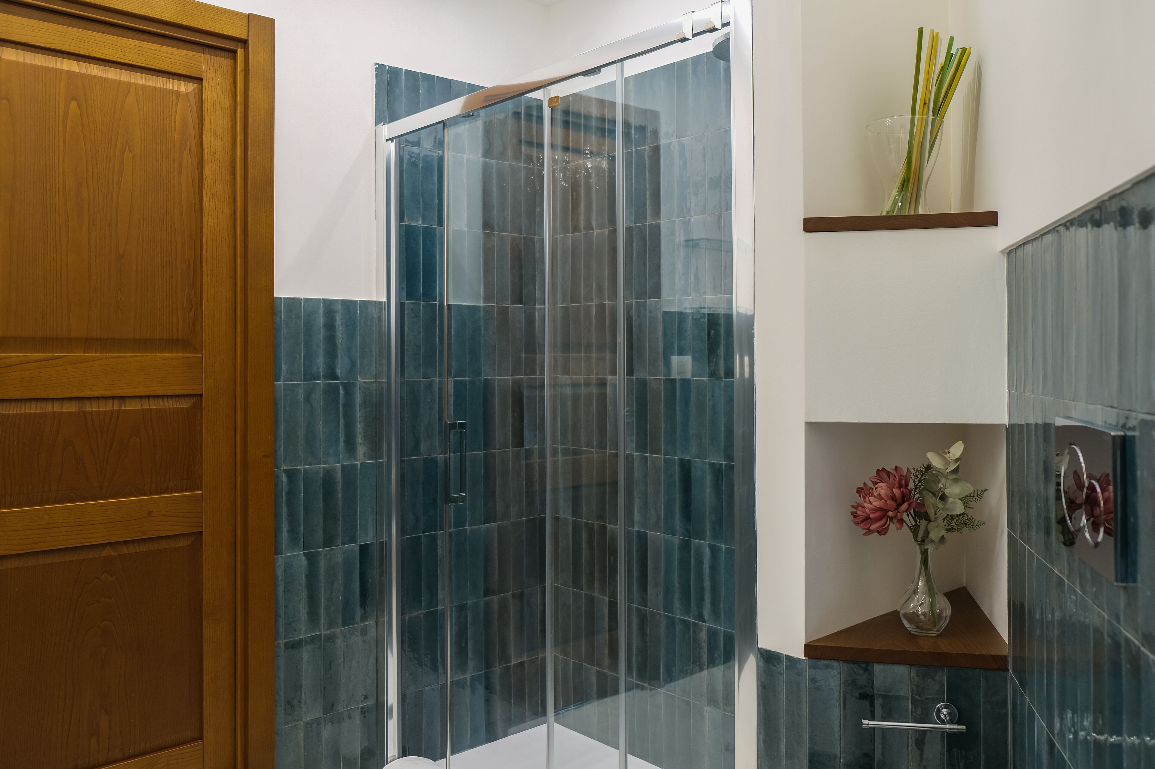 A modern bathroom with a glass shower enclosure, a wooden door, and a shelf with a vase of flowers.