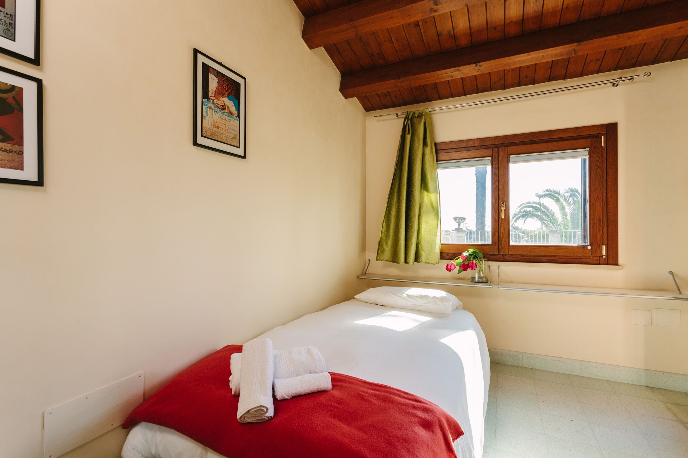 Ein Einzelbett mit roter Decke und weißen Kissen, ein Fenster mit grünem Vorhang und ein an der Wand befestigtes Regal mit einer Blumenvase.