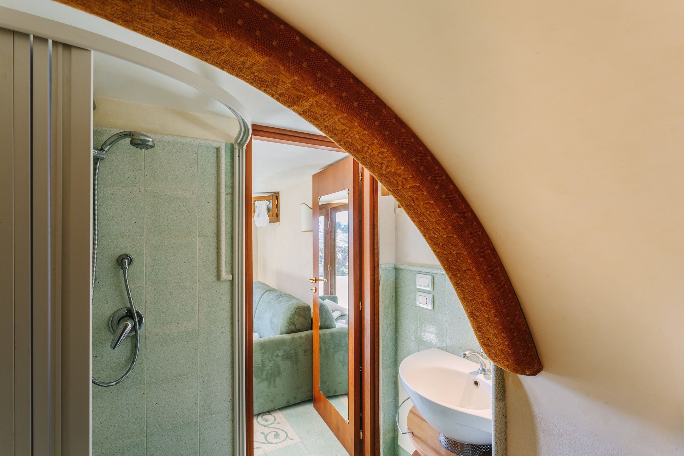 A bathroom with a shower, a sink, and a view of a bedroom through an archway.