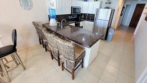 A compact kitchen with a granite countertop, white cabinets, and a built-in refrigerator. There are four woven chairs at the bar area.