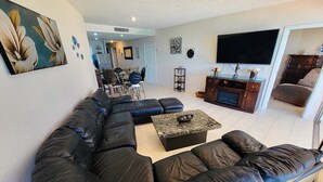 A living room with a black leather sofa, a coffee table, and a TV on a wooden cabinet. There is a dining area in the background with a table and chairs.
