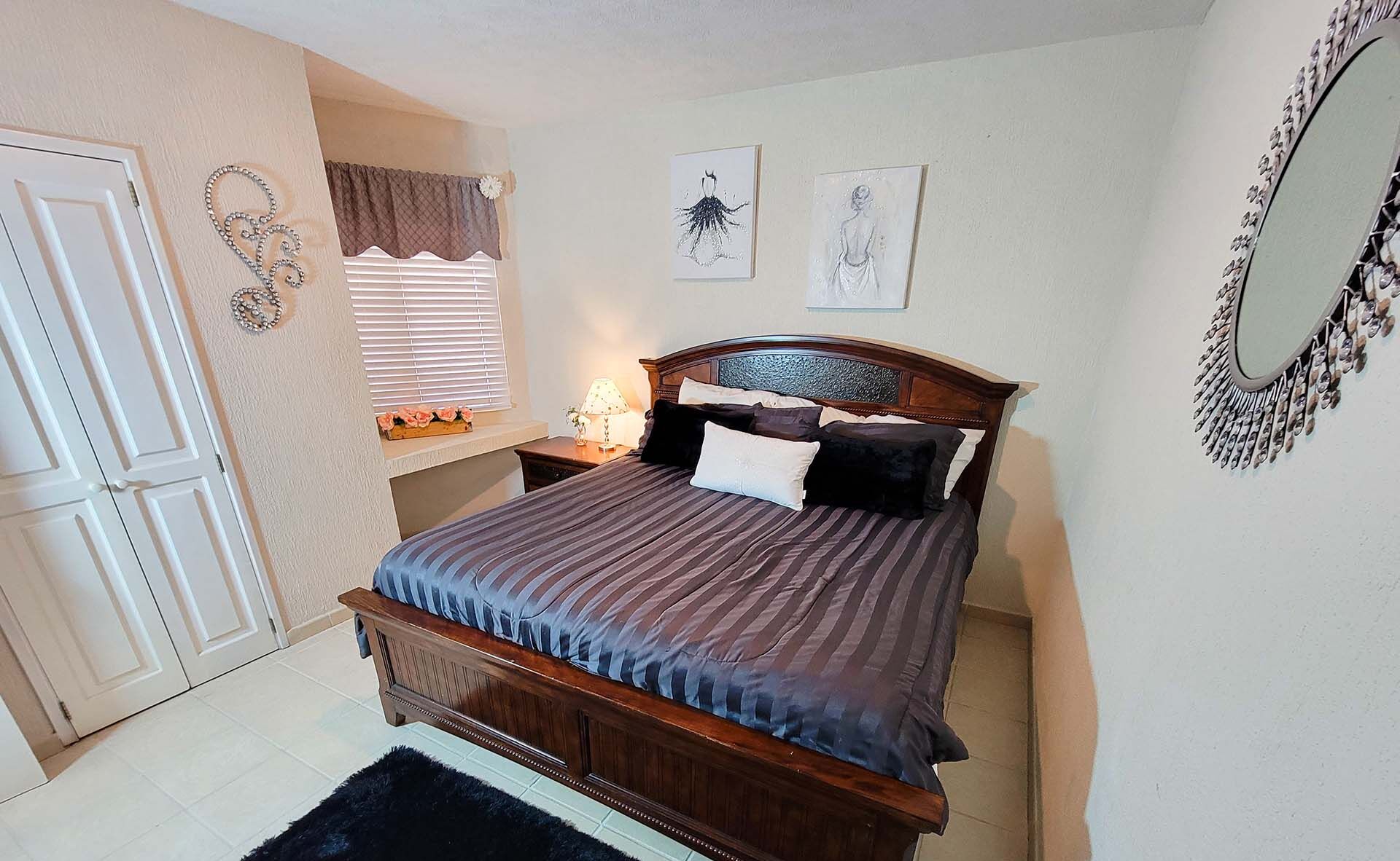 A bedroom with a wooden bed, a nightstand with a lamp, a window with blinds, and a mirror on the wall.
