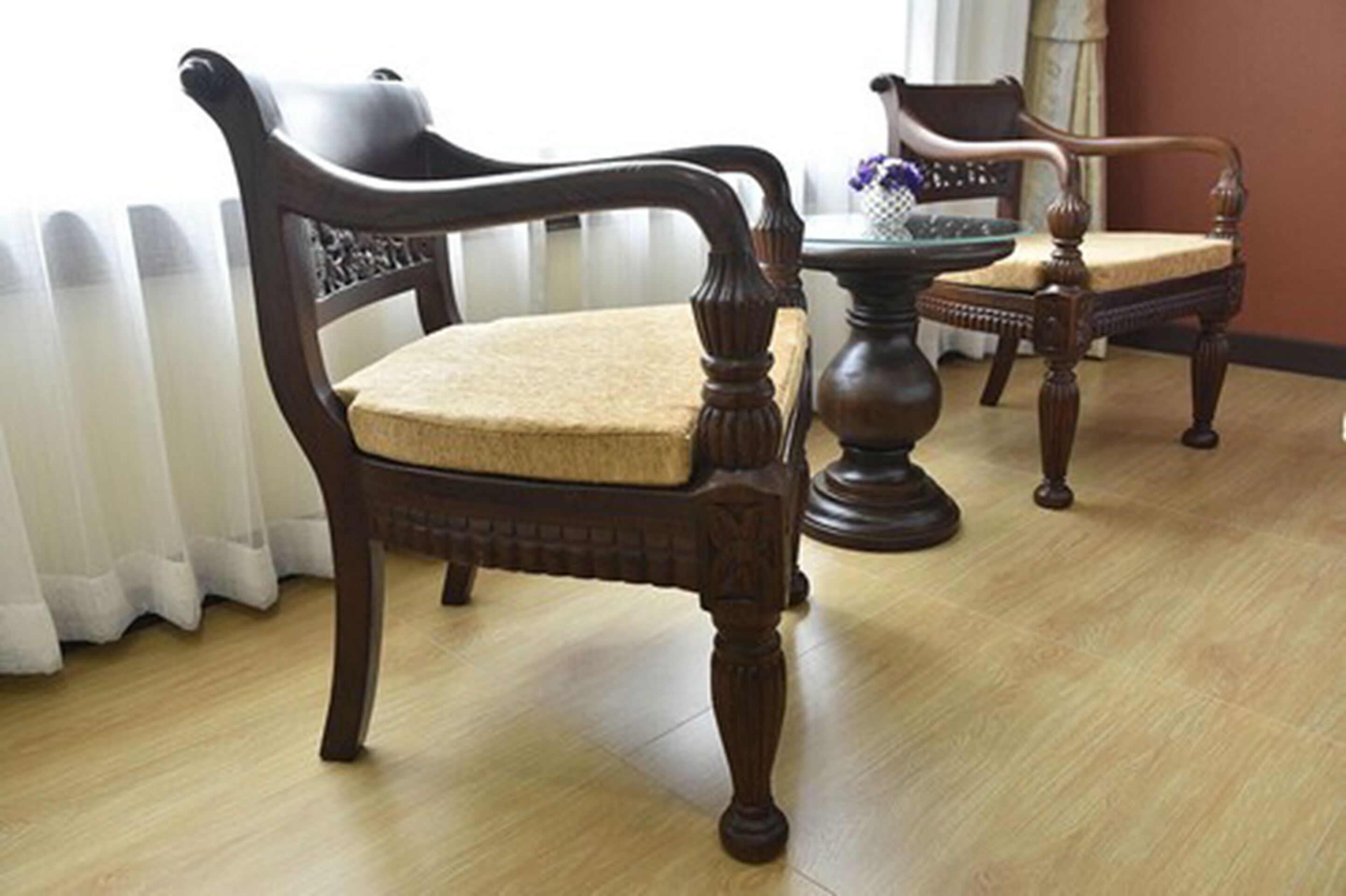 A room with two wooden chairs with cushioned seats and backs, a round wooden table, and a white curtain.