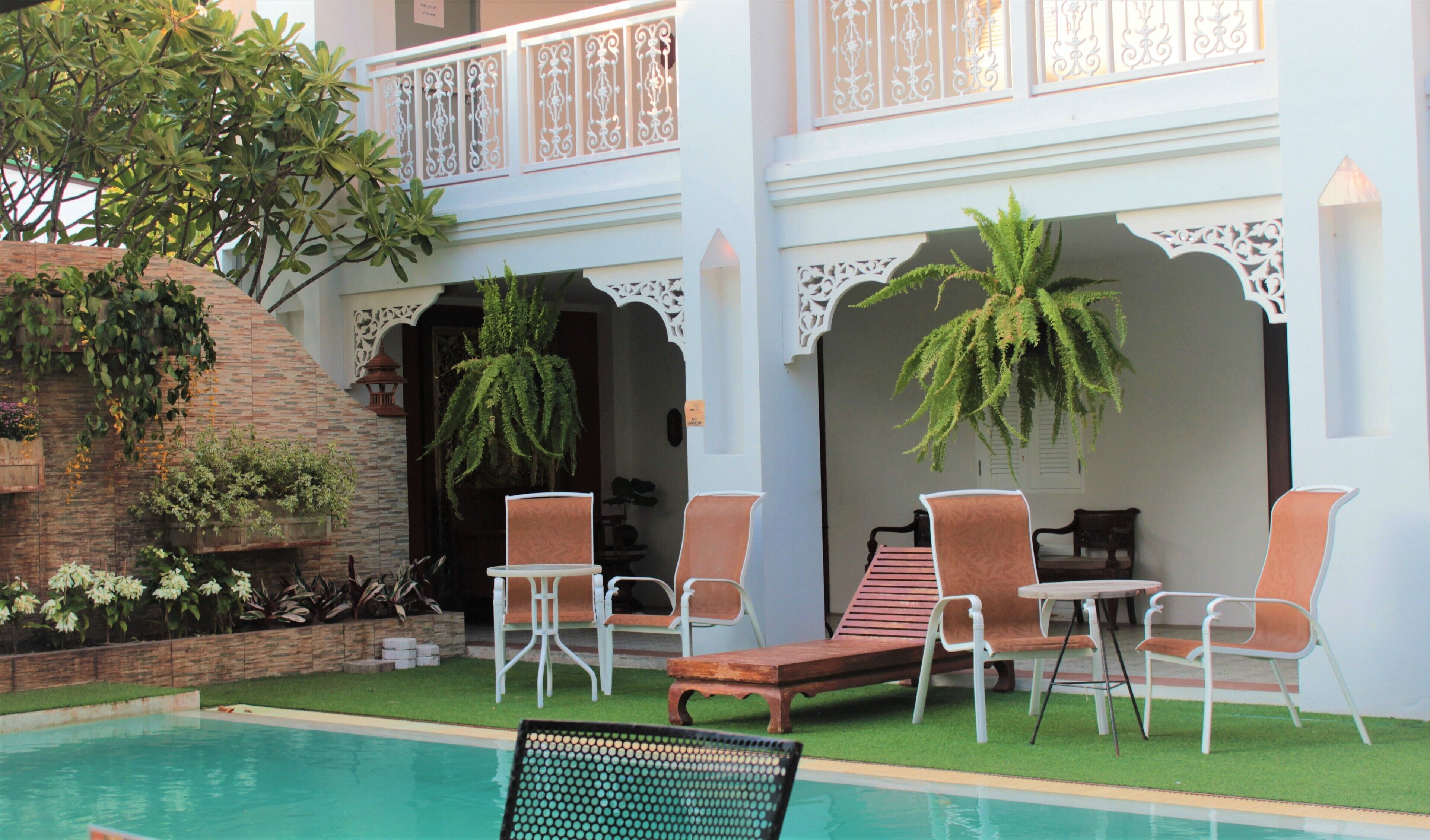Zona junto a la piscina con sillones de descanso y una mesa, rodeada por un edificio blanco con arcos decorativos y plantas colgantes.