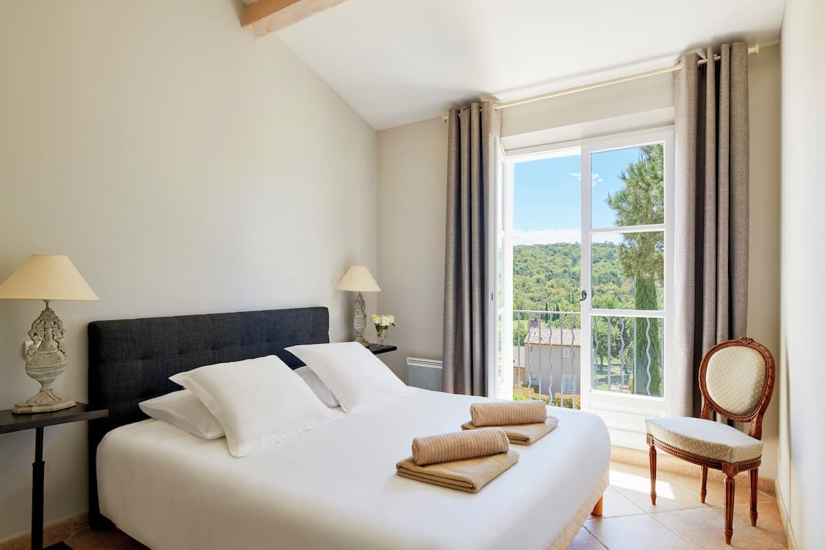 A neatly made bed with white linens and two rolled towels, a wooden nightstand with a lamp, a chair, and a large window with curtains.