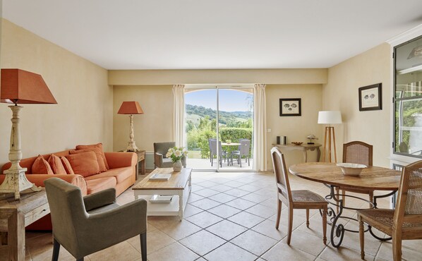 A living room with a sofa, coffee table, and dining table with chairs. There is a view of greenery through a glass door.