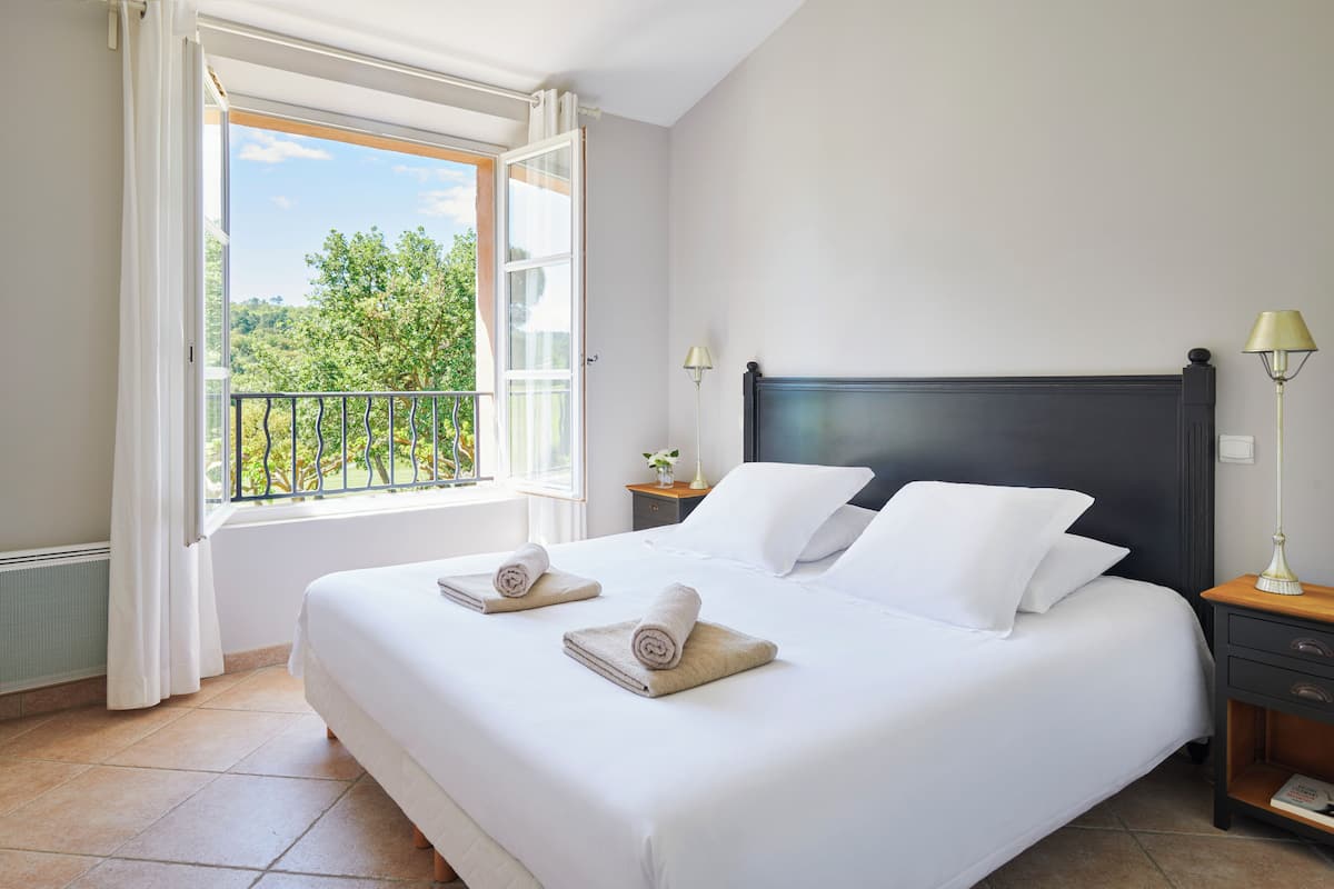 A neatly made bed with white linens and two rolled towels, a bedside lamp, and a view of greenery through an open window.