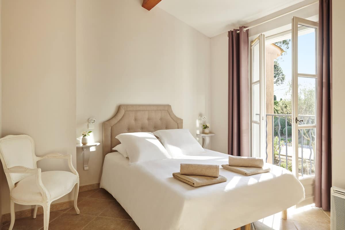 A neatly made bed with two pillows and two folded towels, a white chair, a bedside table with a lamp, and a window with curtains.