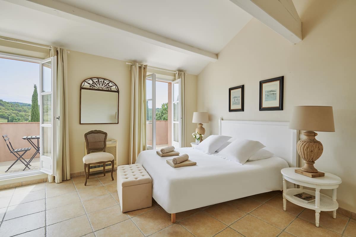 A bedroom with a large bed, a chair, a table with a lamp, and a view of the outdoors.