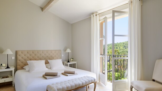 A bedroom with a large bed, a bench, a chair, a nightstand, and a view of the outdoors. - Golf Resort & Country Club Saint-Tropez (Gassin)