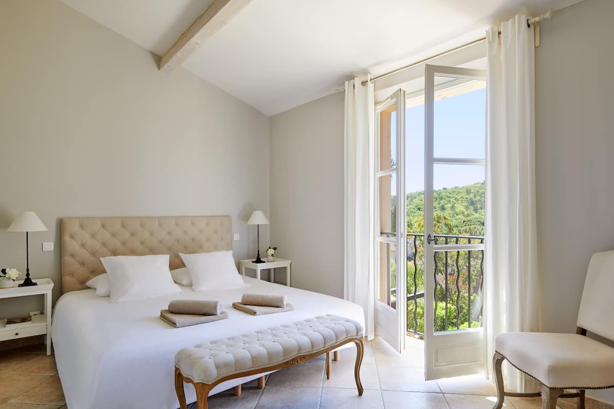 A bedroom with a large bed, a bench, a chair, a nightstand, and a view of the outdoors.