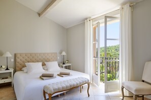 A bedroom with a large bed, a bench, a chair, a nightstand, and a view of the outdoors.