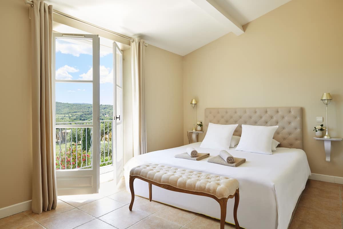 A bedroom with a large bed, a bench, a balcony with a view, and two bedside lamps.