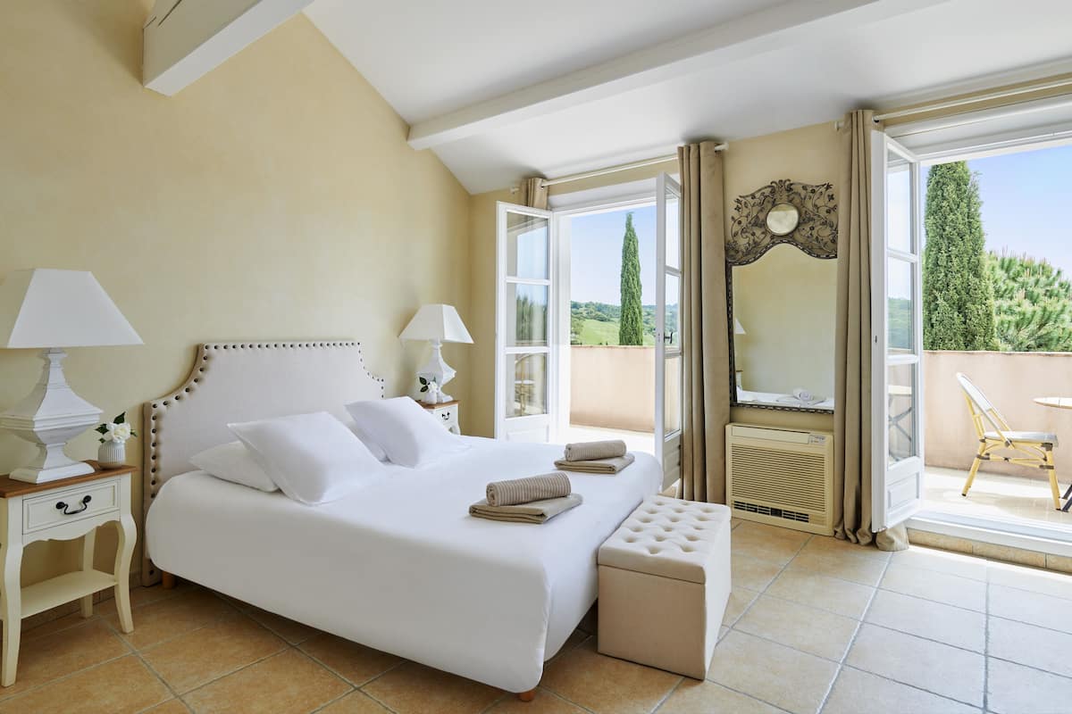 A bedroom with a large bed, two bedside tables with lamps, a bench, and a balcony with a chair.