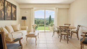 A living room with a sofa, coffee table, and dining table with chairs. There is a view of a garden and hills in the background. - Golf Resort & Country Club Saint-Tropez (Gassin)