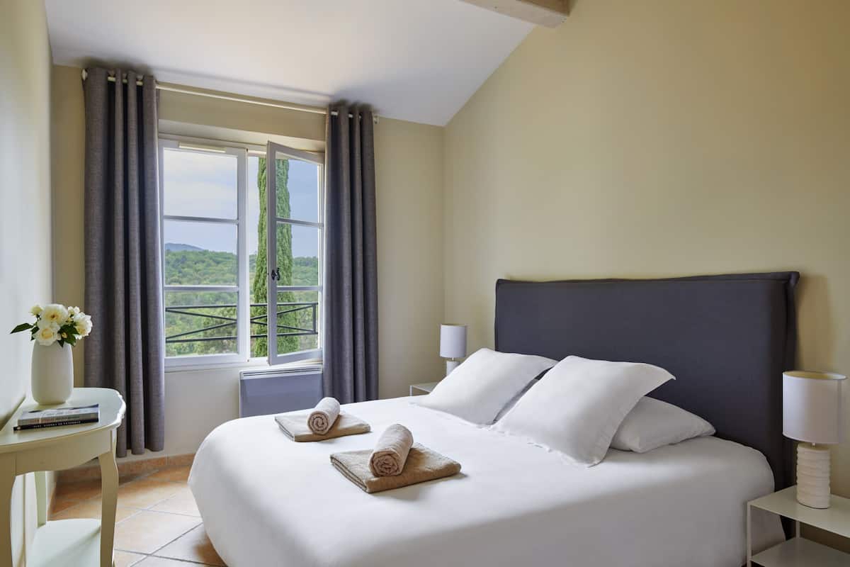 A neatly made bed with white linens and two rolled towels, a bedside table with a lamp, and a window with curtains.