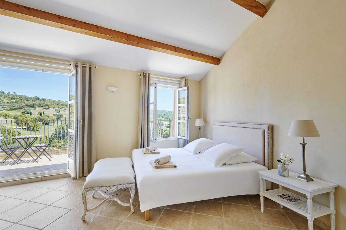 A bedroom with a large bed, a bedside table with a lamp, and a view of a balcony with a table and chairs.