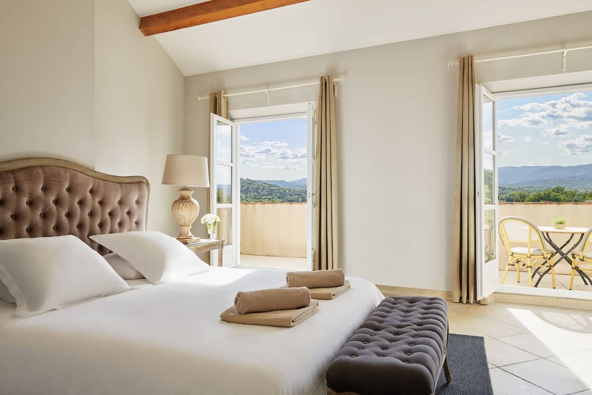 A bedroom with a large bed, a nightstand, a lamp, and a view of a balcony with a table and chairs.